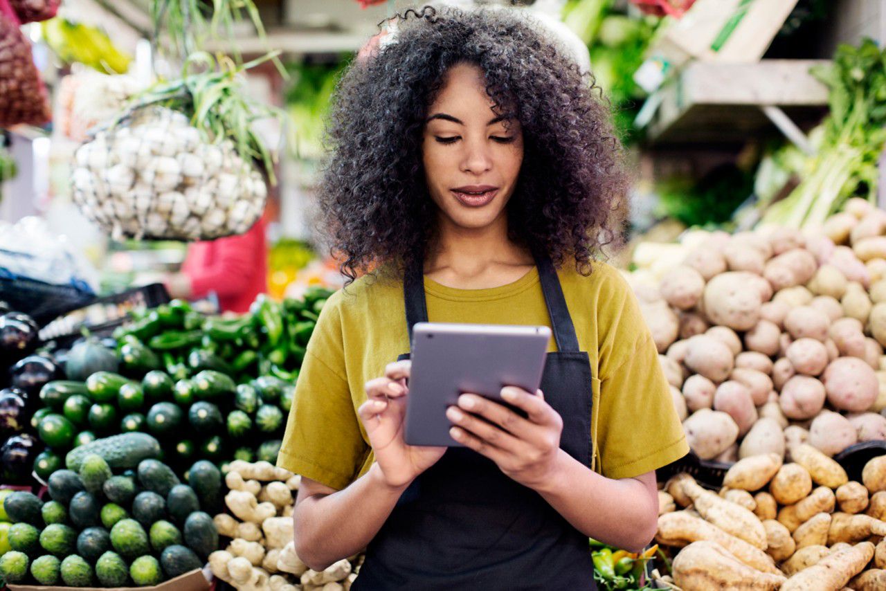 woman wearing apron typing on tablet in front of produce display
